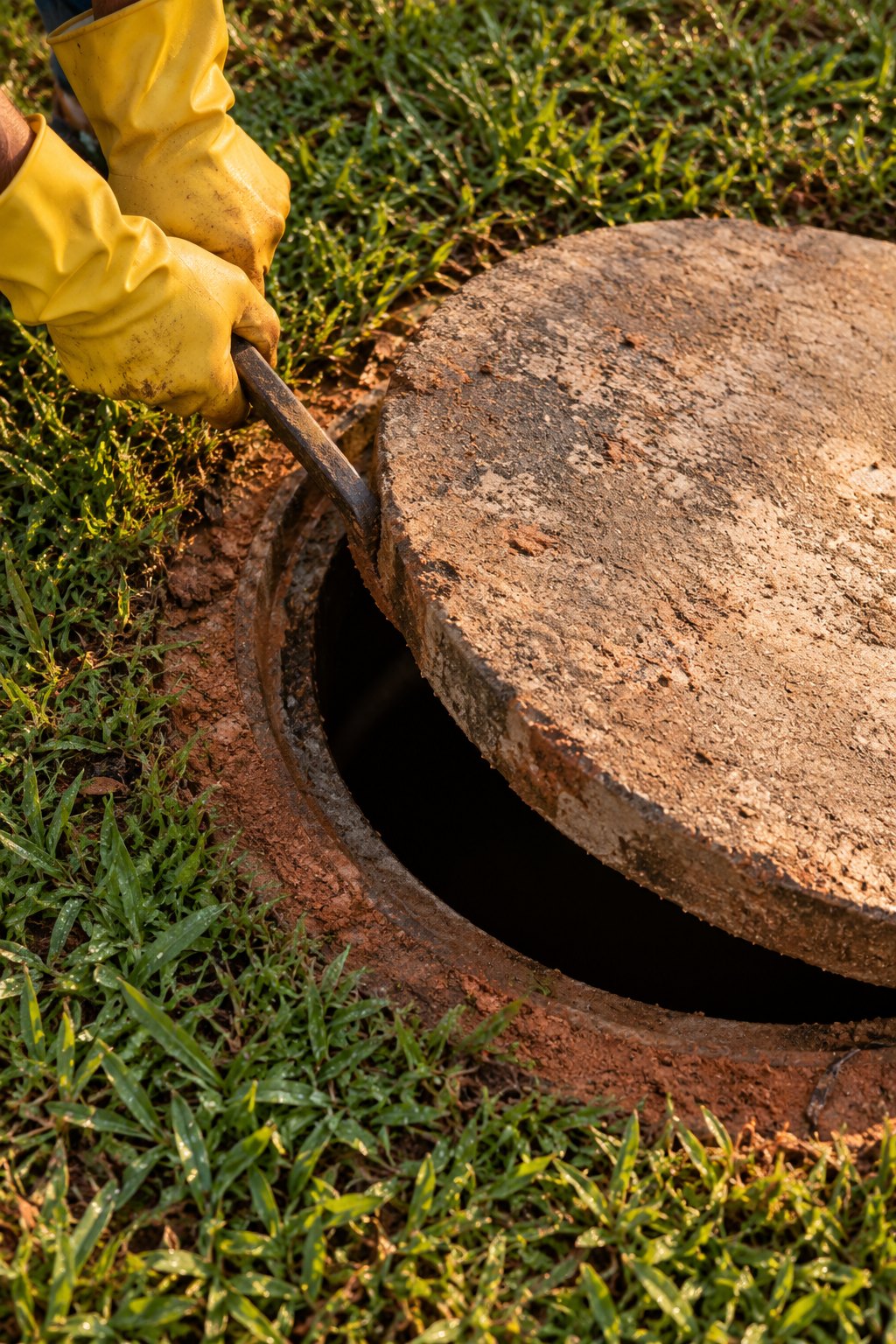 Mãos com luvas usando uma alavanca para erguer a tampa de concreto de uma fossa no gramado